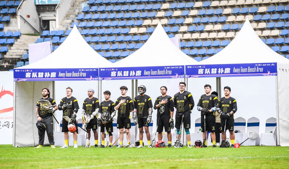 Malaysian lacrosse lining up at sidelines to sing national anthem before face-off.